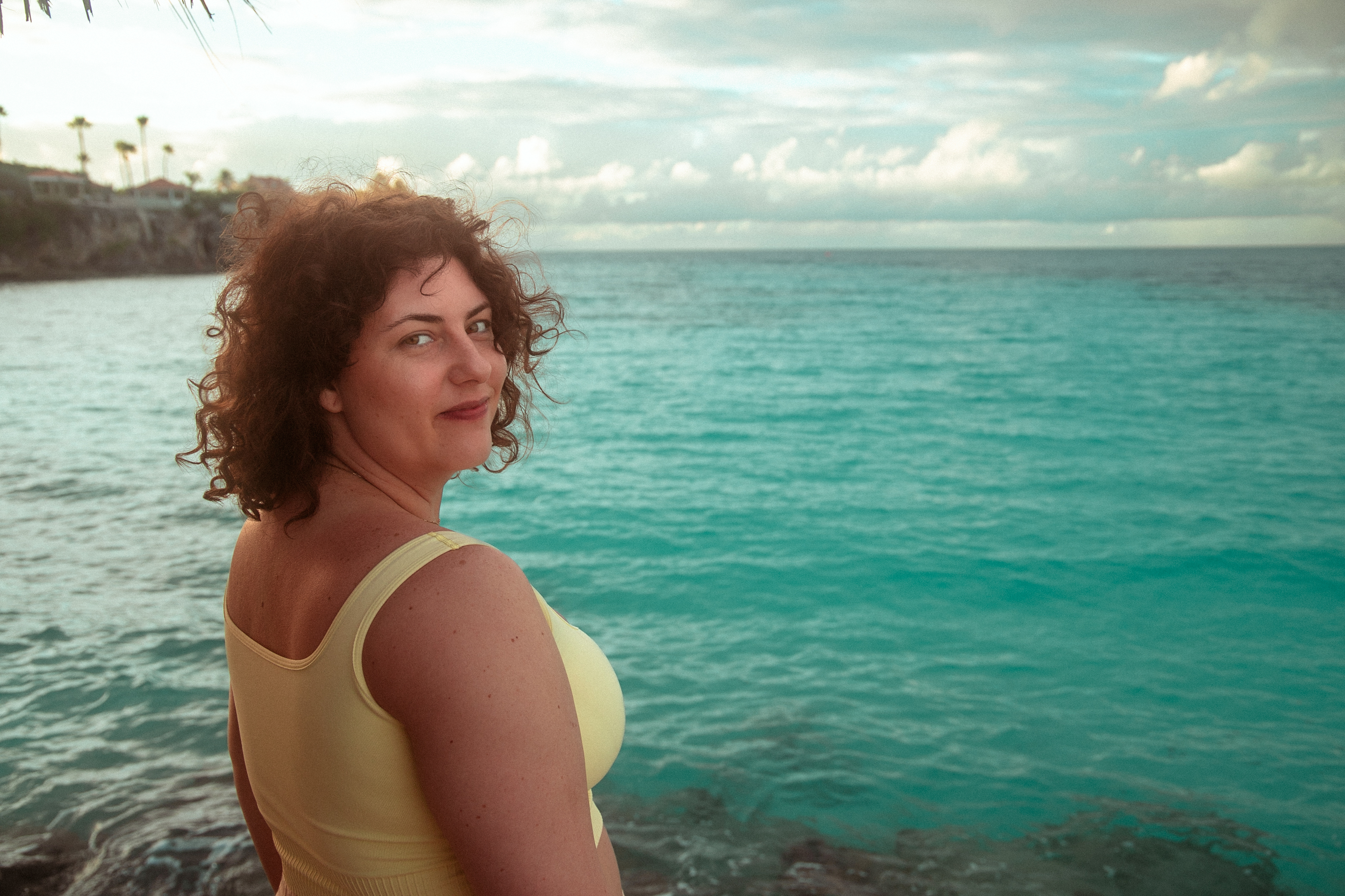 A woman with curly hair and a yellow tank top stands near the ocean, turning slightly to look over her shoulder toward the camera. The water behind her is calm and turquoise, with distant cliffs and a cloudy sky on the horizon. It's the banner image for Only Thing Travel.